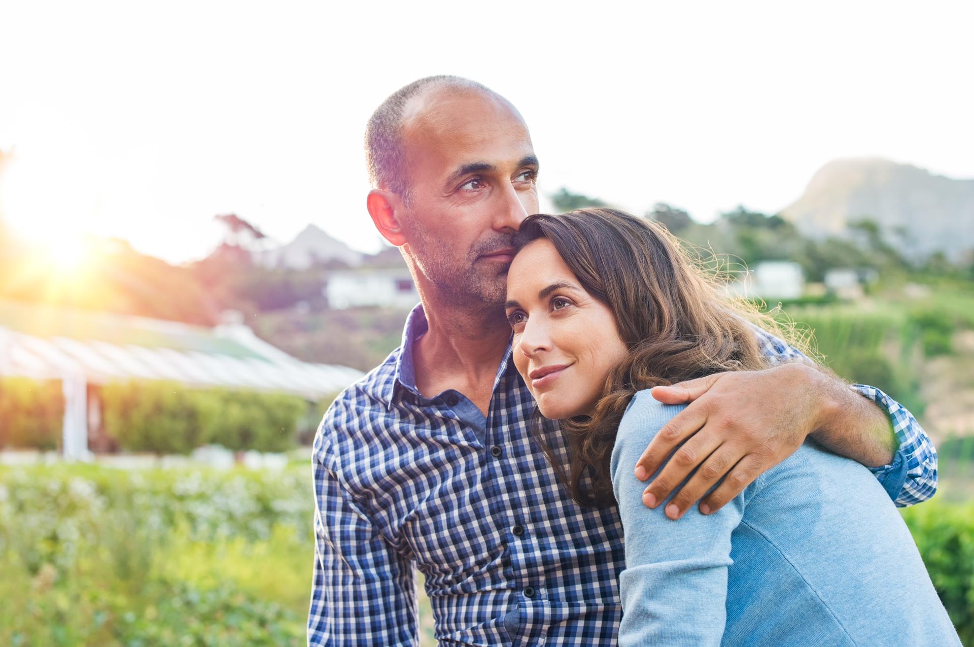 A couple embraces lovingly in a sunlit field, with greenhouses and mountains in the background. The mood is serene and content, capturing a peaceful moment.