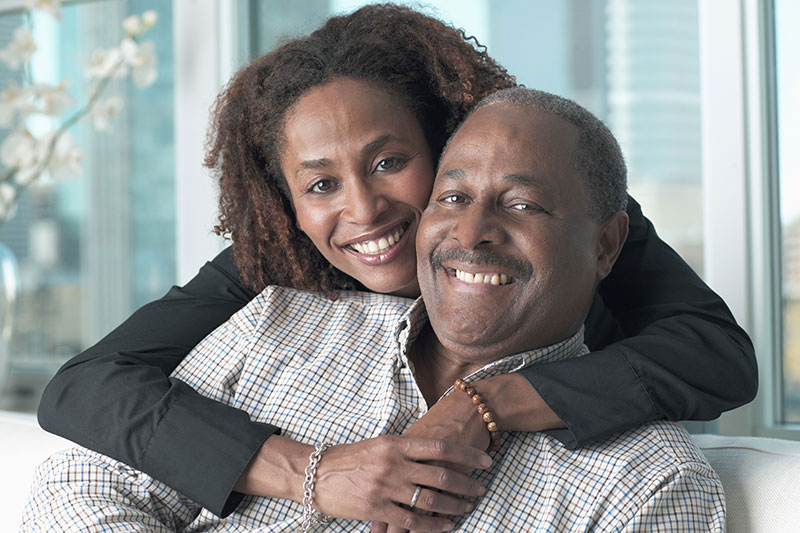 A joyful couple, with the woman embracing the man from behind. They both smile warmly, sitting in a bright room with a city view.