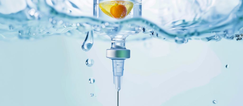 Close-up of an IV drip beneath rippling water with a vibrant orange droplet inside. The scene conveys a clinical yet fresh and rejuvenating feel.